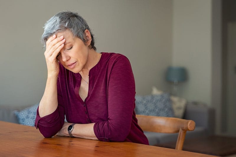 An older woman sits at a table with her head in her hand, clearly in need of tips for stress management for seniors.